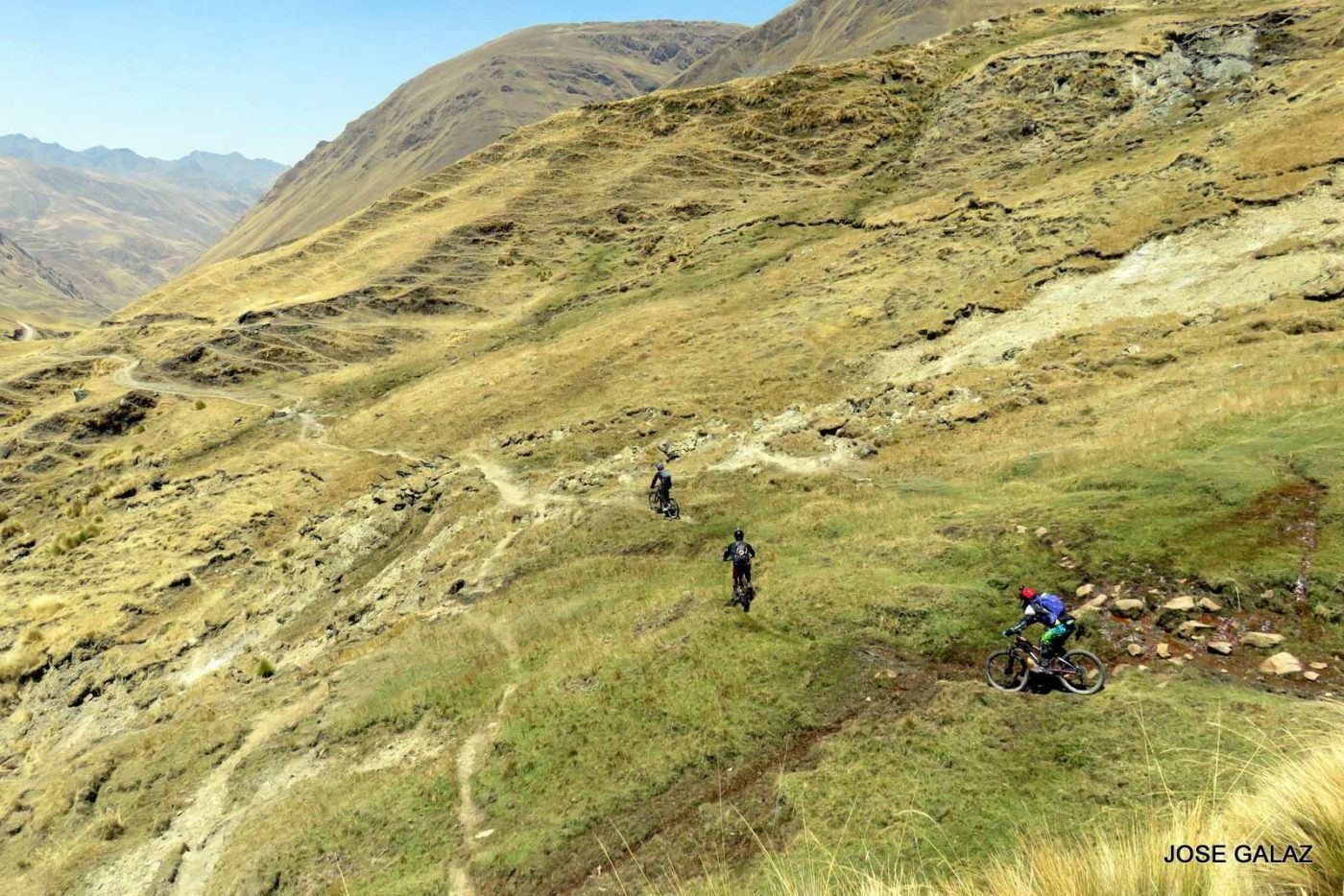 Distant view of three mountain bikers cutting across a grassy mountainside on narrow trails