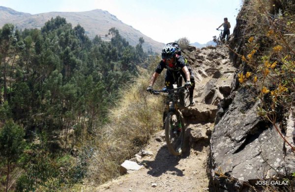 One mountain biker riding quickly down steep rocky trail along cliff's edge while another person walks their bike down behind them