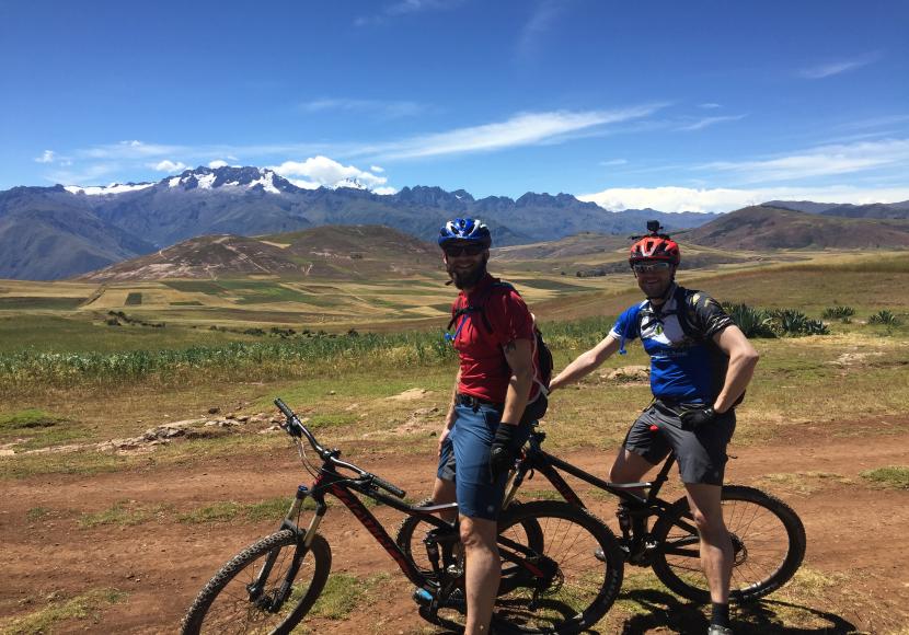 Two men sitting on their mountain bikes on a dirt track in a field in Peru with green hills and mountains in the distance