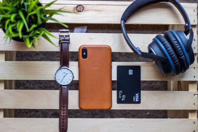 Looking down on items laid out on a wood table: a watch, back of an iPhone leather case, a credit card, and headphones