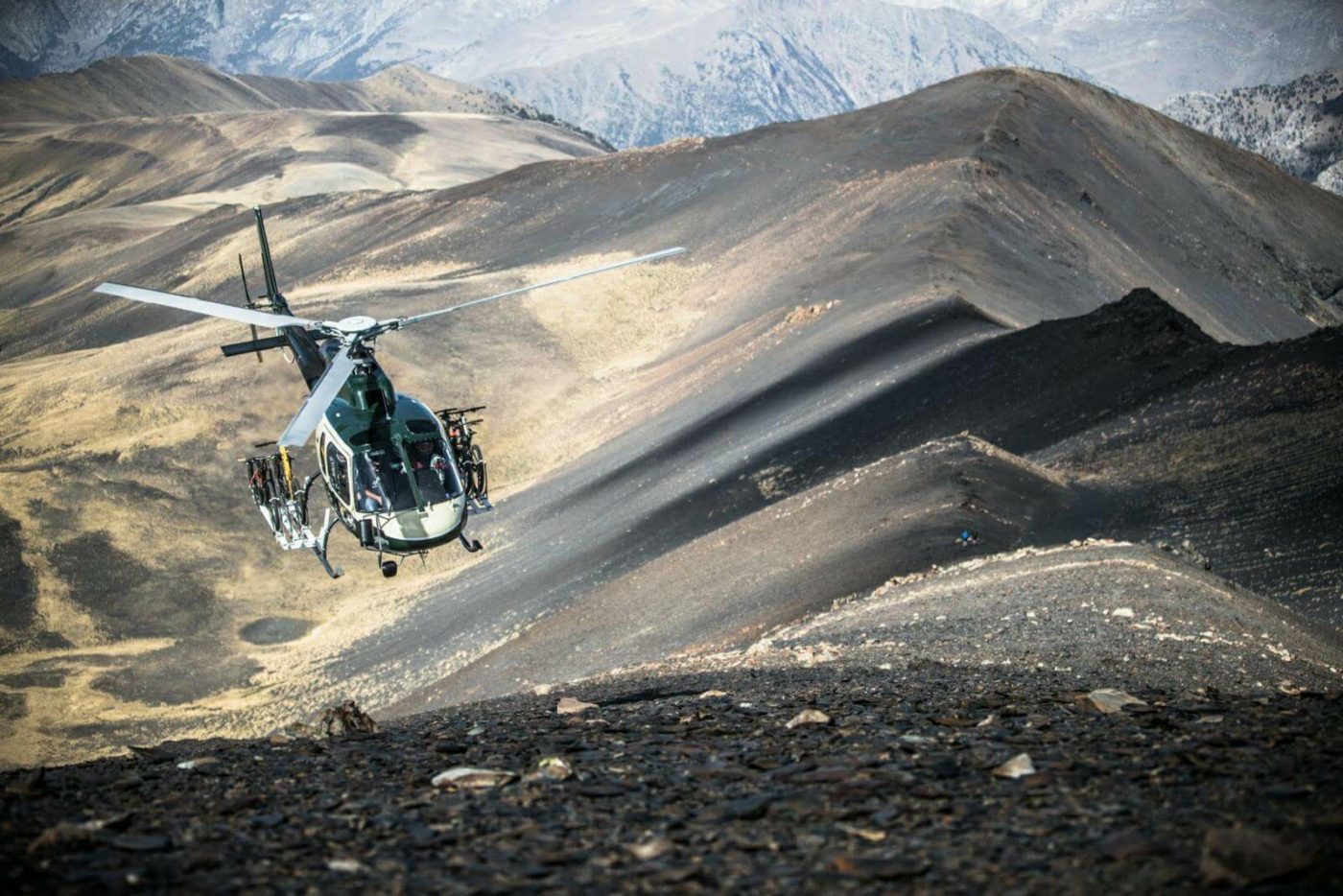 Helicopter carrying mountain bikes coming in to land over mountainous, brown landscape