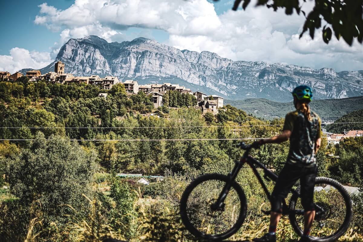 A woman standing next to her mountain bike, looking out over a valley at mountains and the 11th century town of Aínsa, Spain.