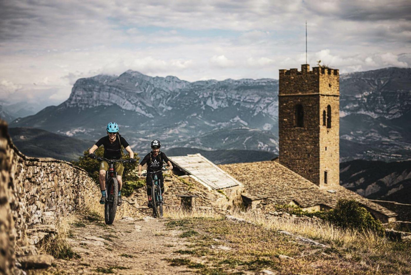 Two mountain bikers riding through Spain, coming up over a hill on a brown dirt path with the Pyrenees and an old stone fortress behind them.