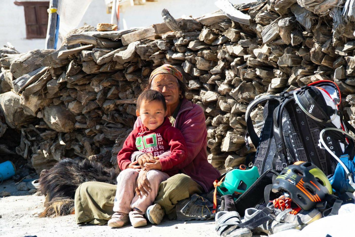 A woman and child in Nepal, sitting on the ground in front of stack of wood as the mountain bike tour passes by