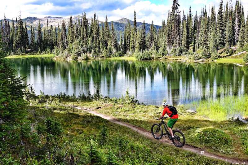 Mountain biker on a trail heading towards a small, glassy lake surrounded by trees and mountains in the distance