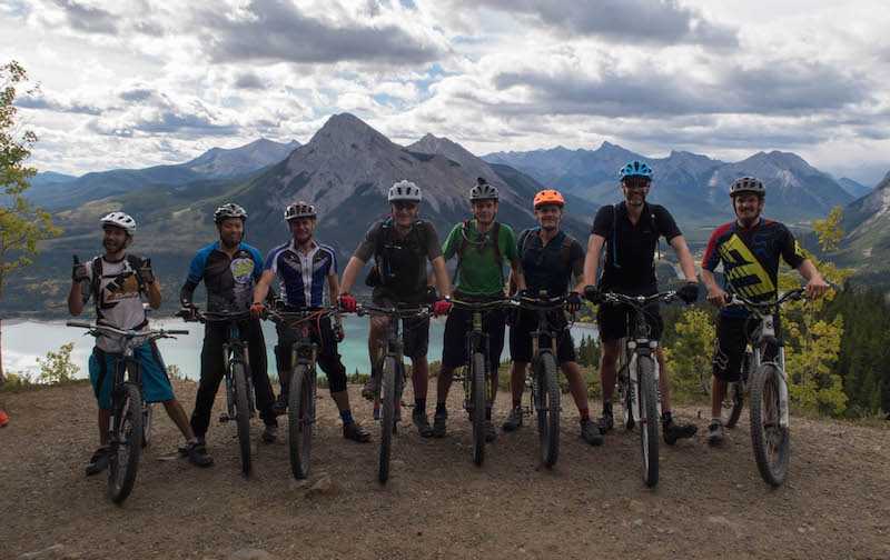 Group of riders posing on their bikes for a group photo with a lake and mountains of British Columbia in the distance