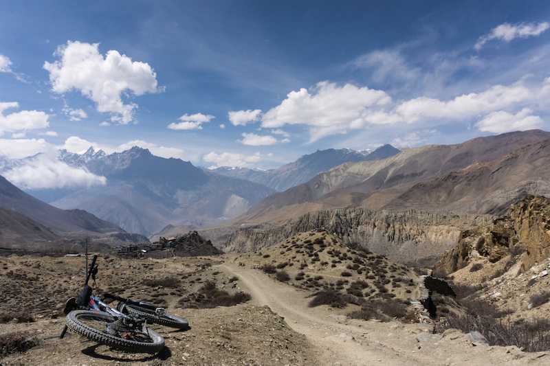 Mountain bike on the ground next to a dusty trail, surrounded by mountains and under blue sky