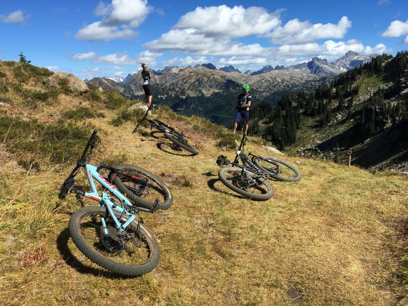Three mountain bikes laying on the ground and two mountain bikers standing and looking out over the mountain valley