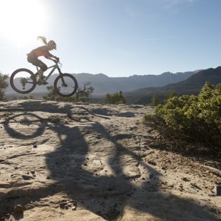 Woman riding mountain bike across desert rock under blue sky and sun