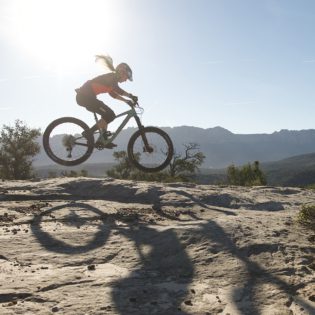 Woman riding mountain bike across desert rock under blue sky and sun