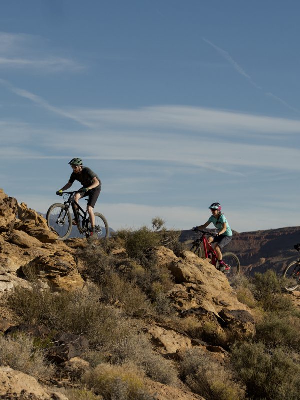 Three people riding mountain bikes over rocky desert terrain in Nevada, with bright blue sky overhead.