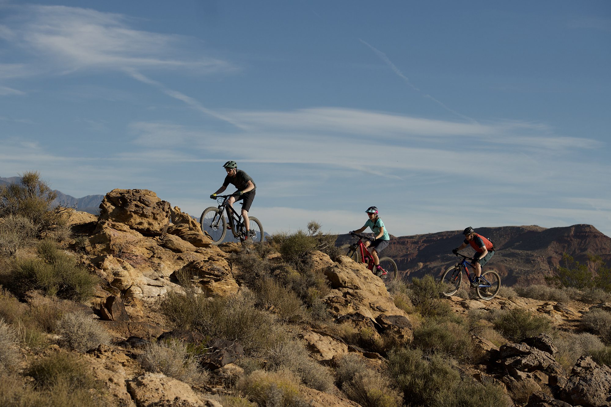 Three people riding mountain bikes over rocky desert terrain in Nevada, with bright blue sky overhead.
