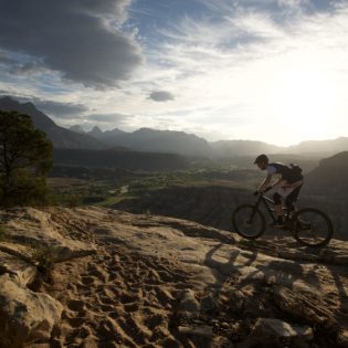 Mountain biker riding across rocky terrain in foreground, with sunsetting over tall rock formations and desert in the background
