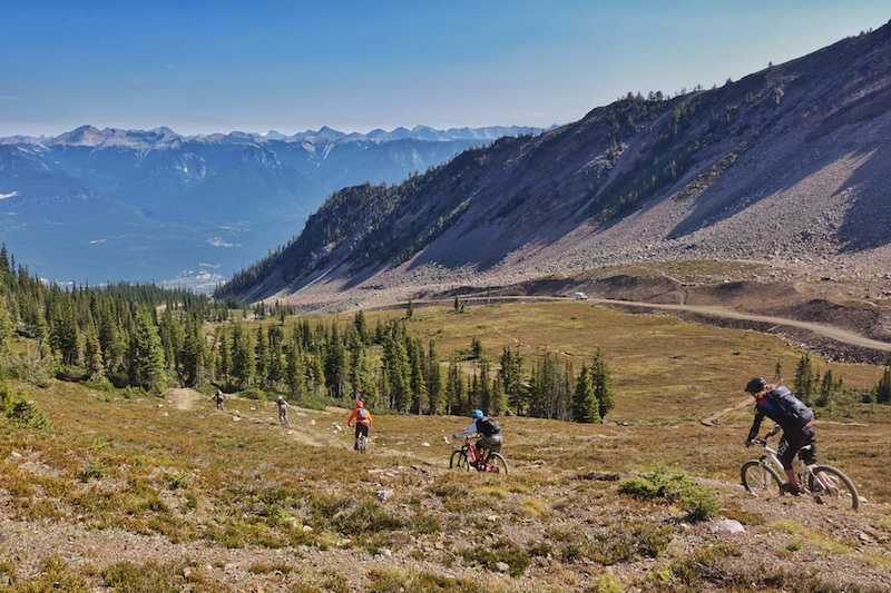 5 Mountain Bikers with lots of space between them riding in a line down a trail in a valley surrounded by mountain
