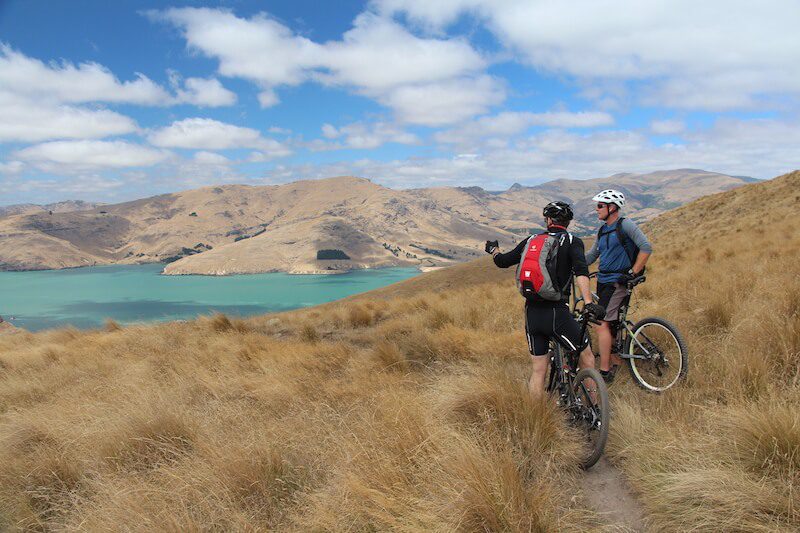 Two mountain bikers stopped to talk in a field of brown grass while looking out over hills and a lake