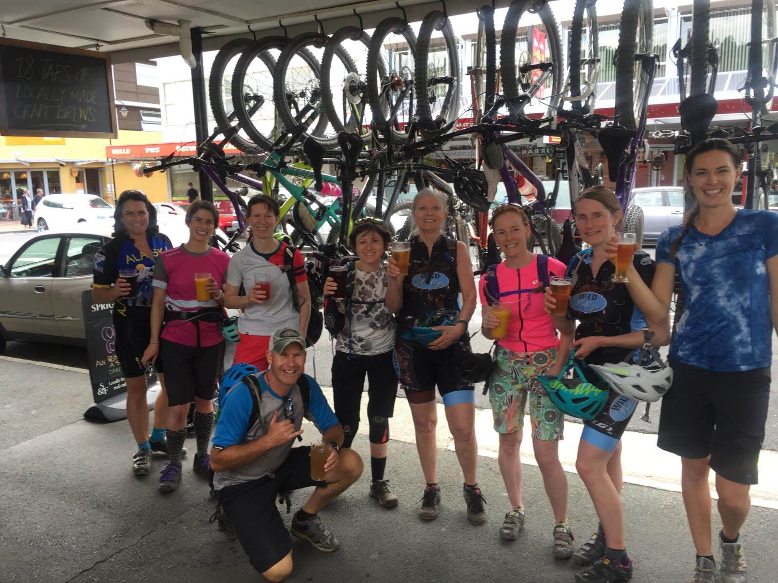 Group of 8 women standing and one man crouching in front of many mountain bikes hanging from ceiling of bike shop