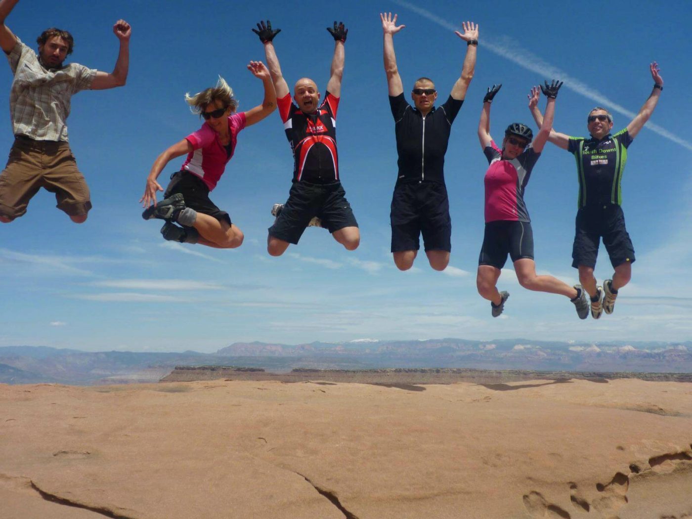 Several people in biking gear leaping into the air and smiling over a reddish brown dry earth landscape in Utah