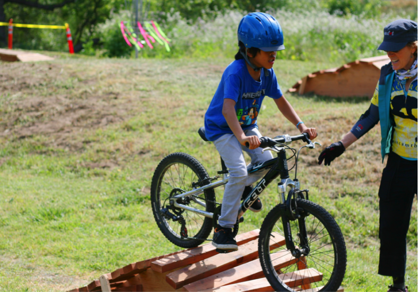 A Kid Around Eleven Years Old Being Coached by an Adult Standing Next to Him While He Learns How to Ride a Mountain Bike Up a Small Ramp