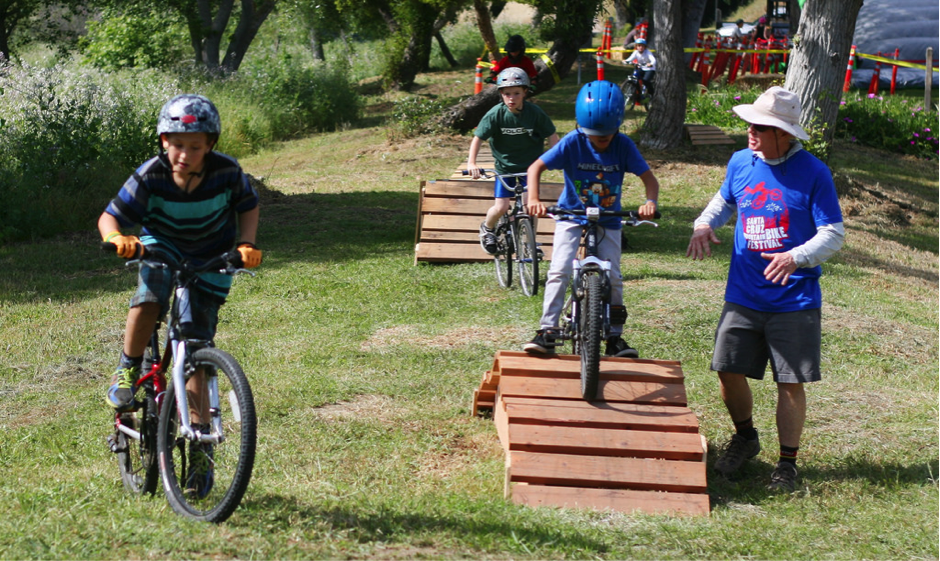 Several Kids Riding Mountain Bikes on Small Ramps in a Field With an Adult Standing Next to One Ramp to Give Them Guidance