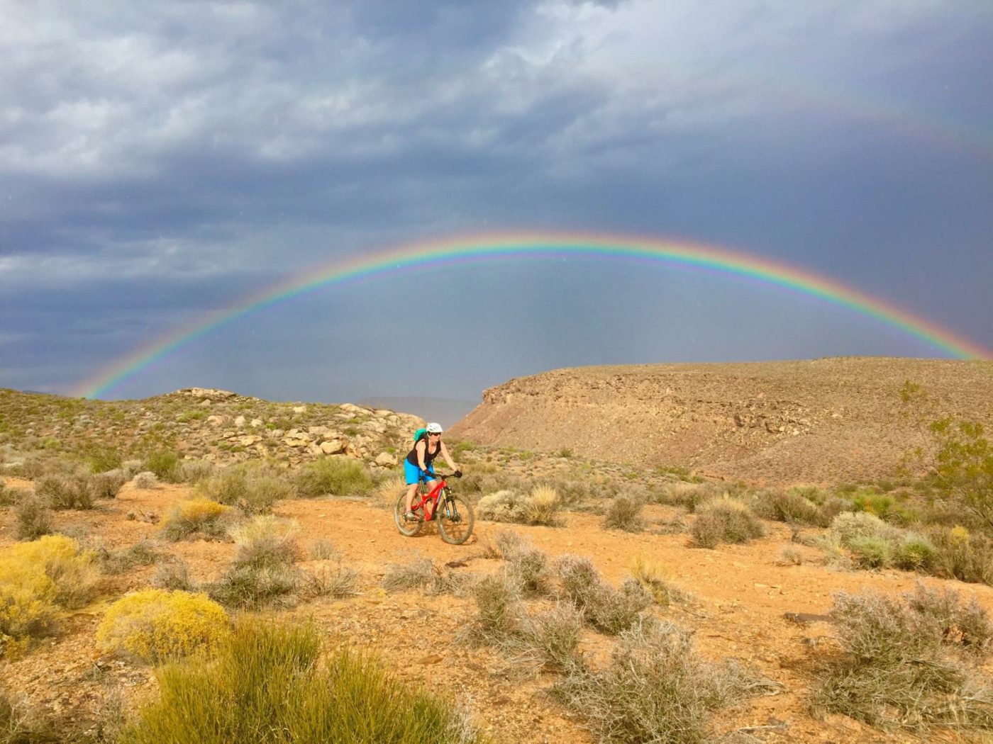Woman riding a mountain bike across red rocky landscape with gray sky and clouds overhead and a rainbow arched over her in the background