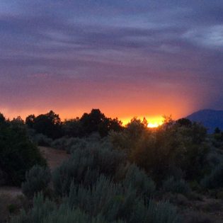 Sun peeking over desert-grass covered hill as it sets under a purplish sky
