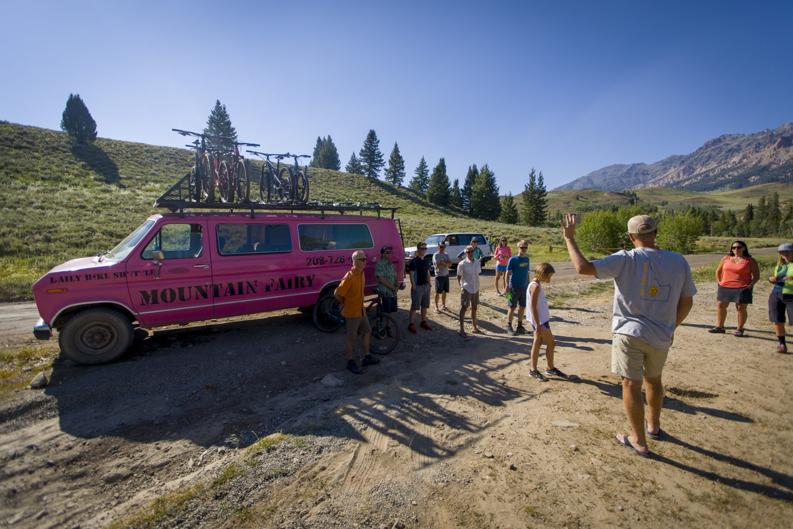Several people gathered in front of a pink van with mountain bike rack on roof, listening to instructions from guide