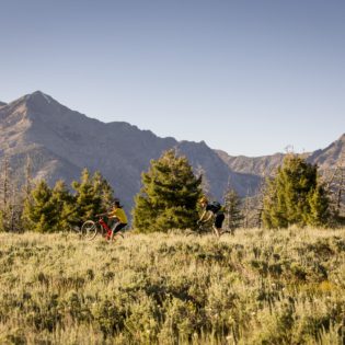 Mountain biker in middle ground riding across grassy field with field in foreground and low mountain peaks in background