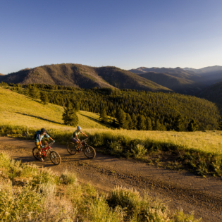 Two mountain bikers riding side by side on wide path surrounded by deep green grass and rolling hillsides