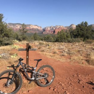 Mountain bike parked in middle of red dirt trail crossing with small wooden cross-shaped sign in middle