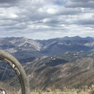 Looking out over rocky mountain valley in distance with a mountain bike tire cutting across the lower right corner in foreground