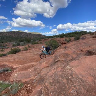 Biker on blue mountain bike riding down red boulders surrounded by desert with blue sky overhead
